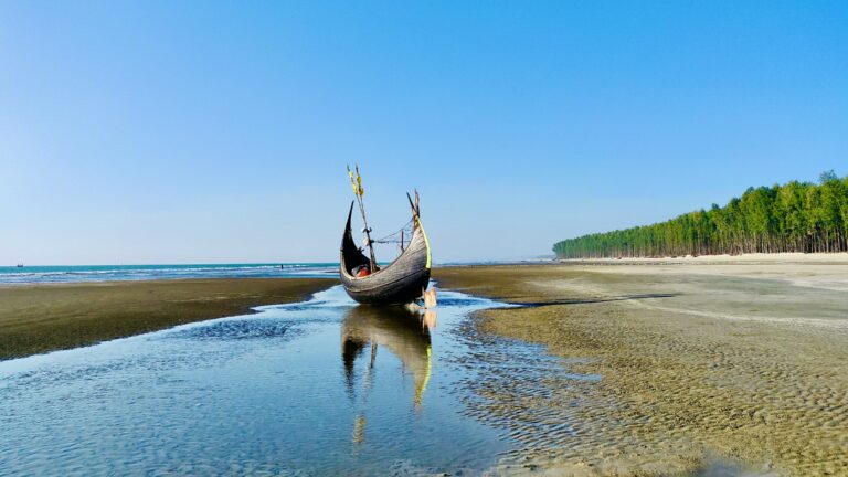 Plaja Cox's Bazar din Bangladesh cu o barcă tradițională și o pădure de pini în fundal.