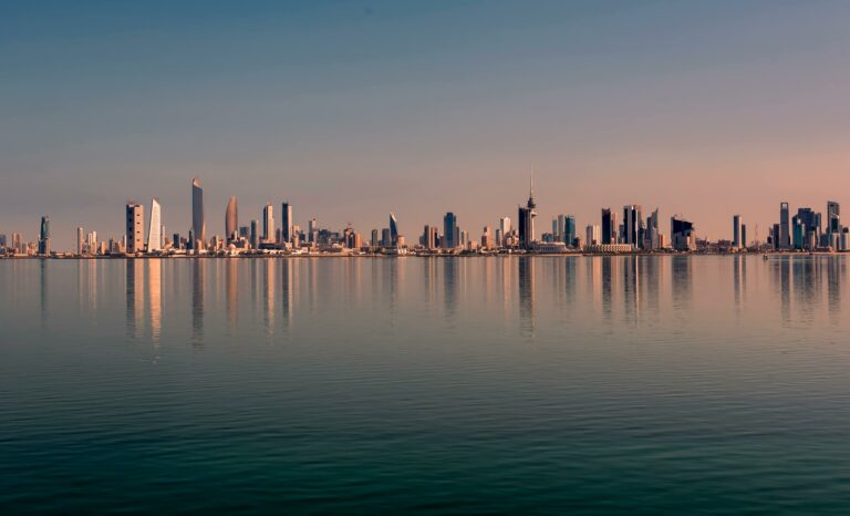 Kuwait City skyline with tall buildings reflected in water at sunset.