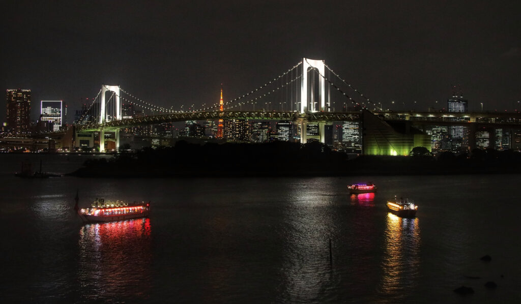 Podul Rainbow iluminat noaptea în Odaiba, Tokyo, cu barci luminoase pe apă și Tokyo Tower vizibilă în fundal.
