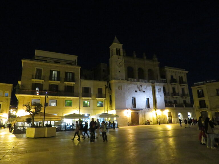 Piazza Mercantile din Bari noaptea, cu clădiri istorice luminate și oameni care se plimbă.