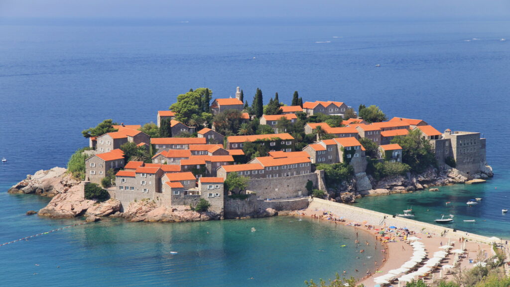 Vedere panoramică a insulei Sveti Stefan, stațiune de lux situată în Budva, Muntenegru, cu acoperișuri roșii și plajă spectaculoasă.