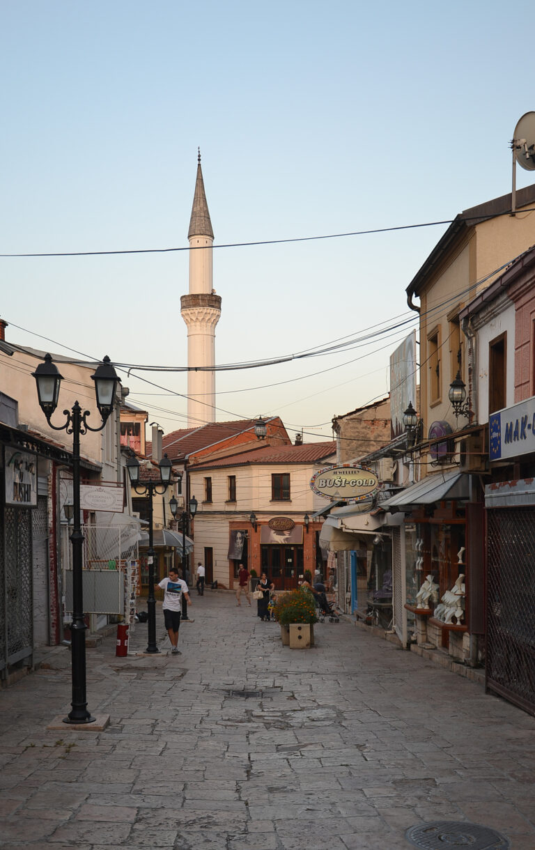 Strada nel Vecchio Bazaar di Skopje con un'atmosfera balcanica tradizionale e un minareto visibile sullo sfondo.