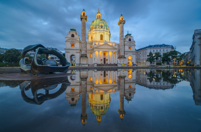 La Chiesa di San Carlo a Vienna, Austria, riflessa nell'acqua sotto un cielo blu al tramonto, con la sua architettura illuminata e suggestiva.