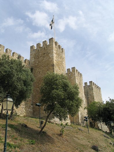 Castello di Sao Jorge con torri di pietra e una bandiera che sventola al vento, fotografato da un angolo inferiore.