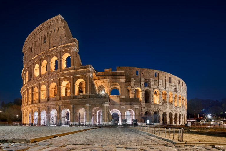 Colosseumul luminat nocturn la Roma, arhitectura iconică dezvăluită sub cerul întunecat.
