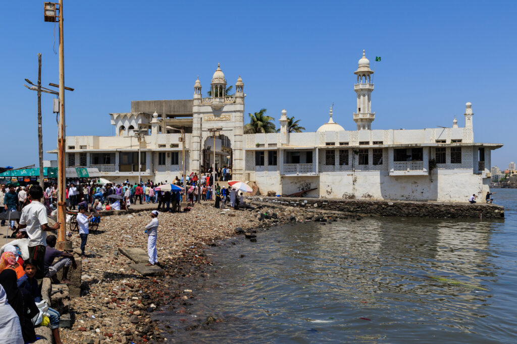 Mausoleo Haji Ali a Mumbai visto dalla riva, con dettagli architettonici e turisti nelle vicinanze.