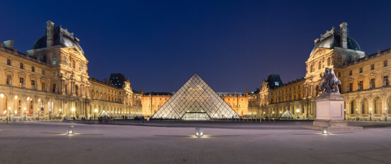 Muzeul Luvru din Paris noaptea, cu piramida de sticlă iluminată și arhitectura clasică în fundal.