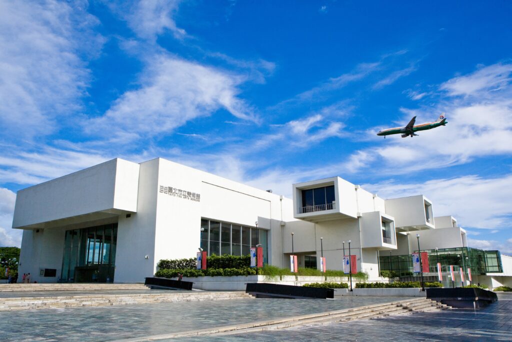 Vista frontale dell'edificio del Taipei Fine Arts Museum sotto un cielo azzurro brillante, con un aereo che vola sopra.