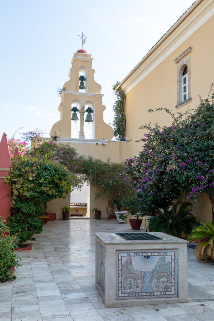 Cortile interno del Monastero di Paleokastritsa, Corfù, Grecia, con vegetazione rigogliosa e campane.