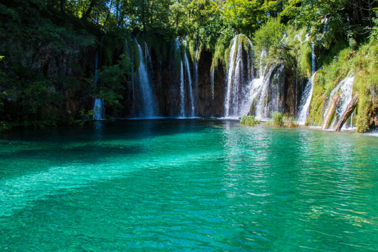 Cascada pitorească din Plitvice National Park reflectată în apă albastră cristalină, înconjurată de vegetație verde.