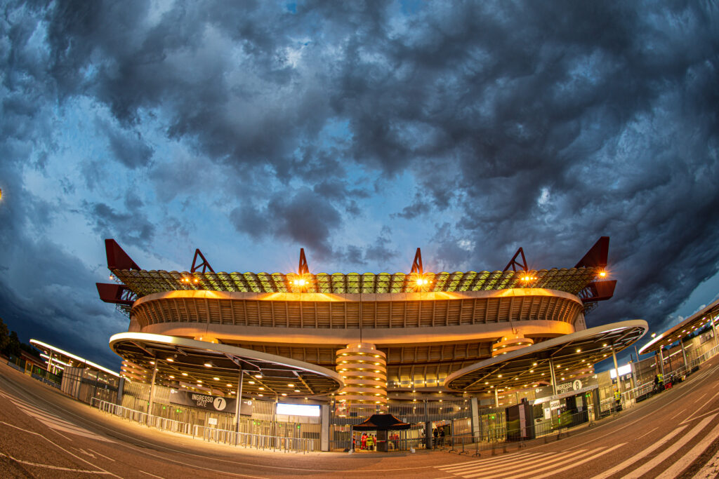 Stadio San Siro di Milano illuminato al tramonto, con nuvole drammatiche sopra.
