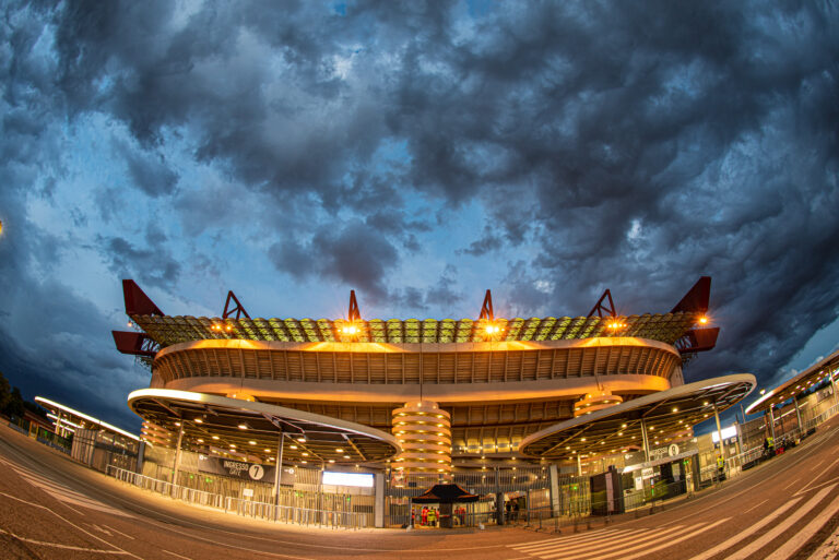 Stadio San Siro di Milano illuminato al tramonto, con nuvole drammatiche sopra.