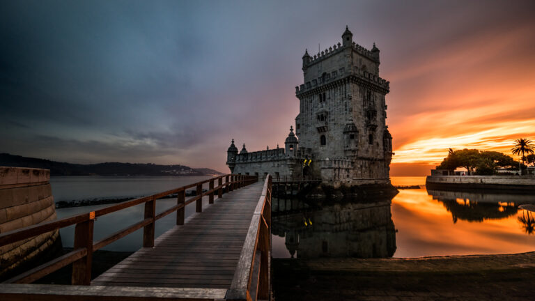 Turnul Belem din Lisabona la apus, cu un pod din lemn și cer senin-portocaliu reflectat în apă.